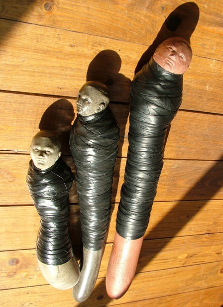 Sculptures de Camille Fonteret : trois formes allongées à visages humains, enrubannées, posées sur un plancher bois (Gembloux, Belgique).