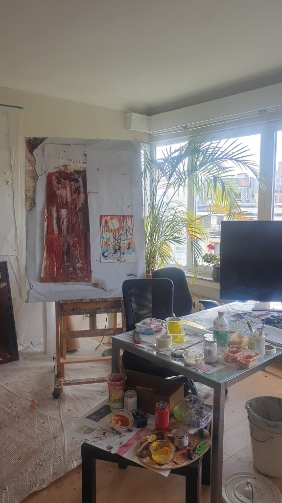 Artist studio of Jérôme Selosse – Expressionist Painter in Brussels: easel with canvas, small pinned painting, glass table with paints and palettes, large window and palm plant (Ixelles).