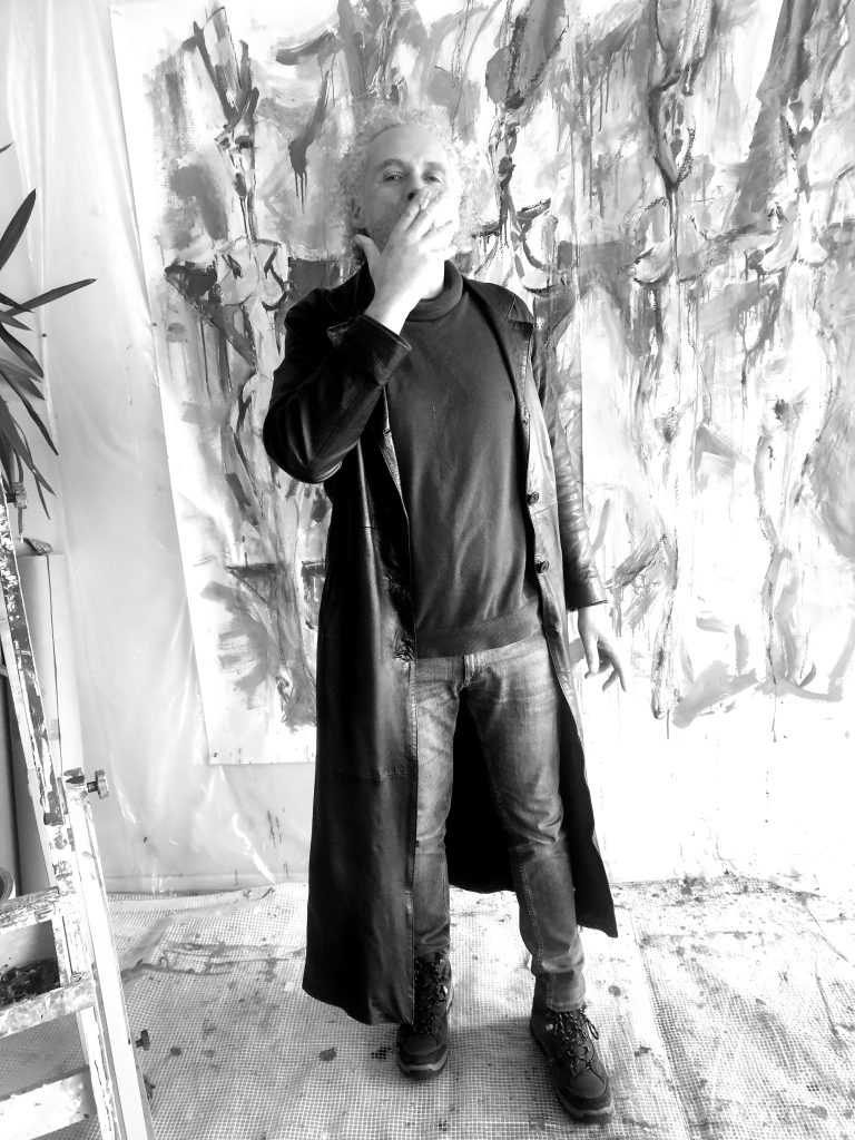 Portrait noir et blanc de l'artiste peintre Jérôme Selosse posant devant une œuvre monumentale sur le Féminin Sacré dans son studio à Bruxelles.