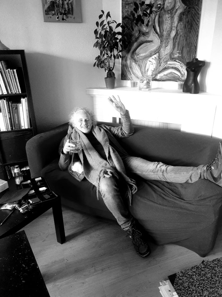 Portrait noir et blanc de l'artiste Jérôme Selosse détendu sur un canapé, saluant l'objectif un verre à la main, entouré de ses livres d'art et peintures.