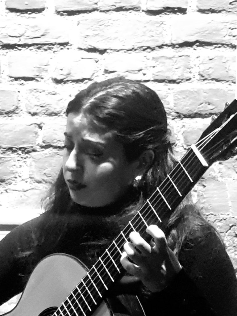 Photographie en noir et blanc de la guitariste Paula Aguilo (Trío Azahar) jouant de la guitare classique. Concert lors d'une exposition d'art à La Cambre. Elle interprète Capricho espagnol devant un mur de briques. Photo par Jérôme Selosse.