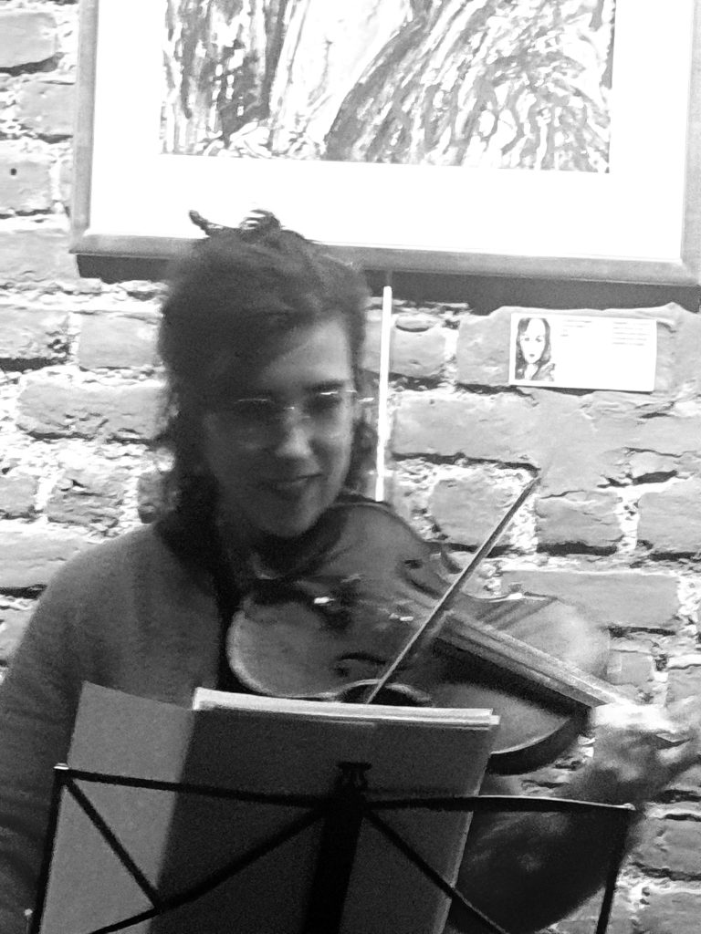 Photographie en noir et blanc de la violoniste Cristina Mura jouant du violon. La musicienne du Trío Azahar lit sa partition sur un pupitre lors de l'interprétation du Capricho espagnol, devant un mur de briques exposant des œuvres d'art. Photographie Jérôme Selosse. Cambre Expo
