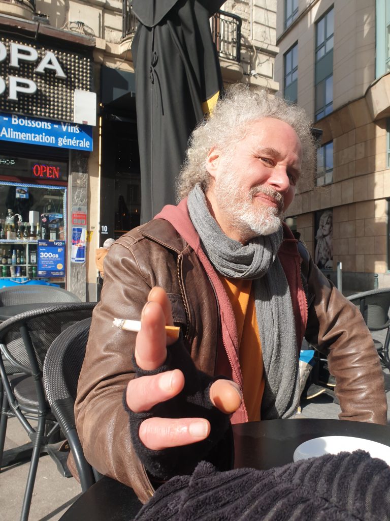 Portrait de l'artiste peintre expressionniste Jérôme Selosse, souriant à la terrasse d'un café. Une scène de vie authentique et ensoleillée au cœur de Bruxelles.