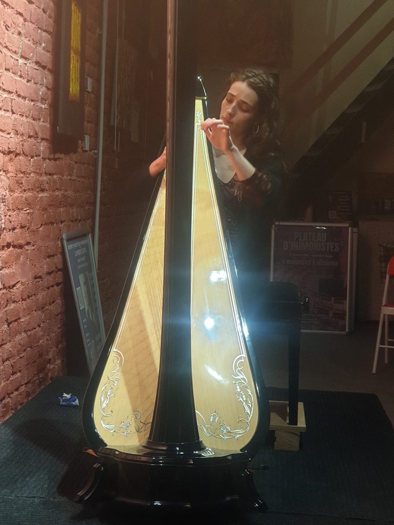 Photographie de la harpiste Louise Fauveau-Sellin jouant de la harpe de concert à Cambre Expo, Bruxelles. Ambiance feutrée devant un mur de briques lors d'un événement musique et peinture.