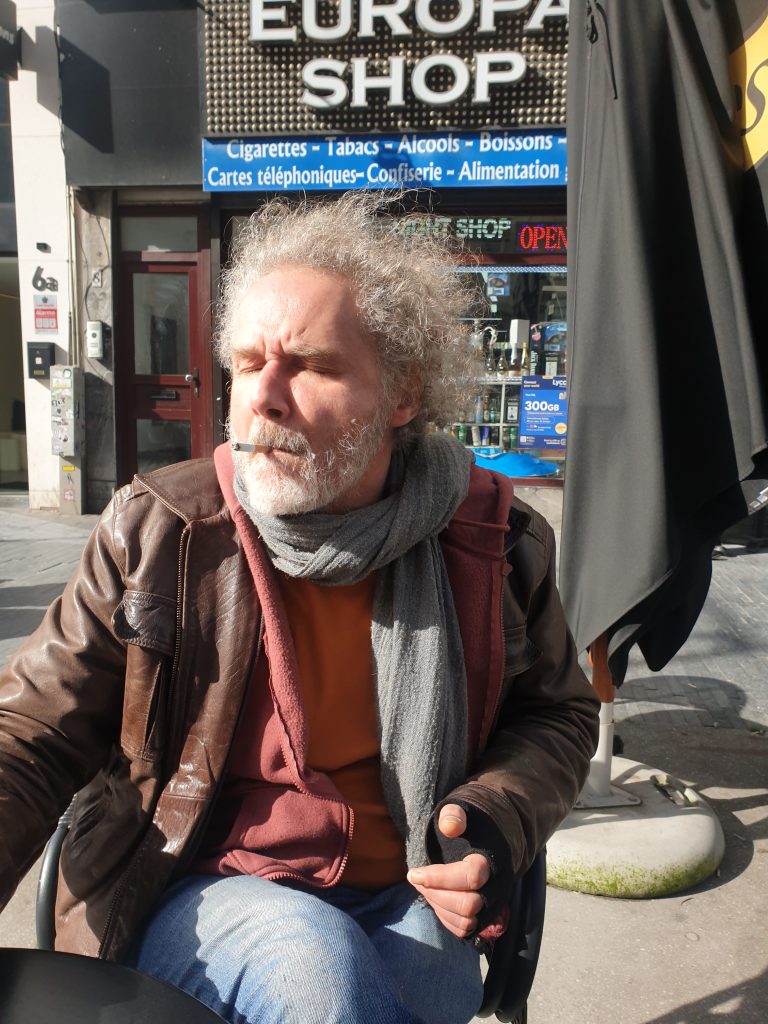 Portrait ensoleillé de l'artiste peintre expressionniste Jérôme Selosse, les yeux fermés, se détendant à la terrasse d'un café dans les rues de Bruxelles.